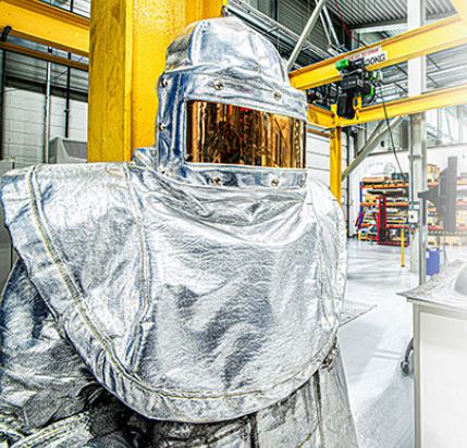 Person standing in manufacturing environment wearing silver boiler suit with orange reflective visor