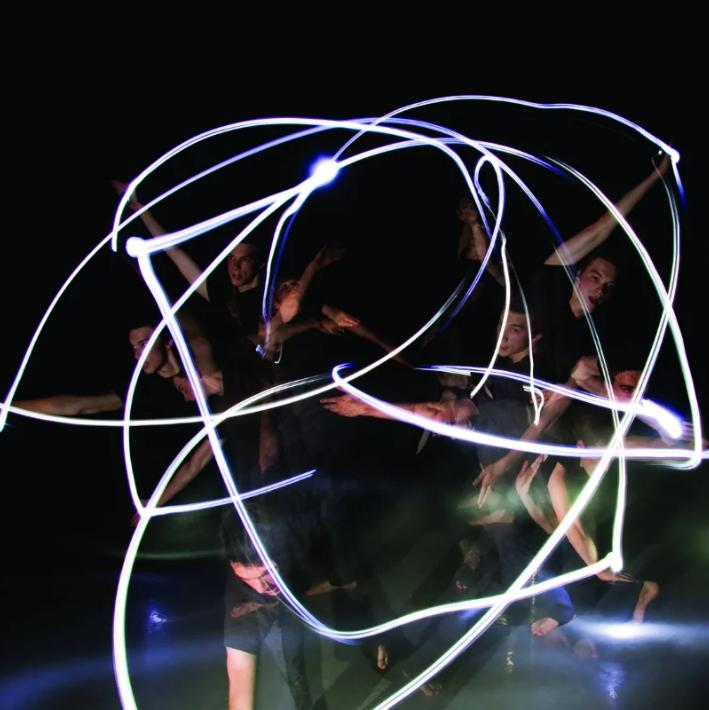 A group of dancers in a dark room with beams of light following their arm movements.