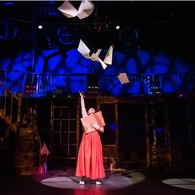 Woman in middle of a dark stage with spotlight on her throwing papers above her head