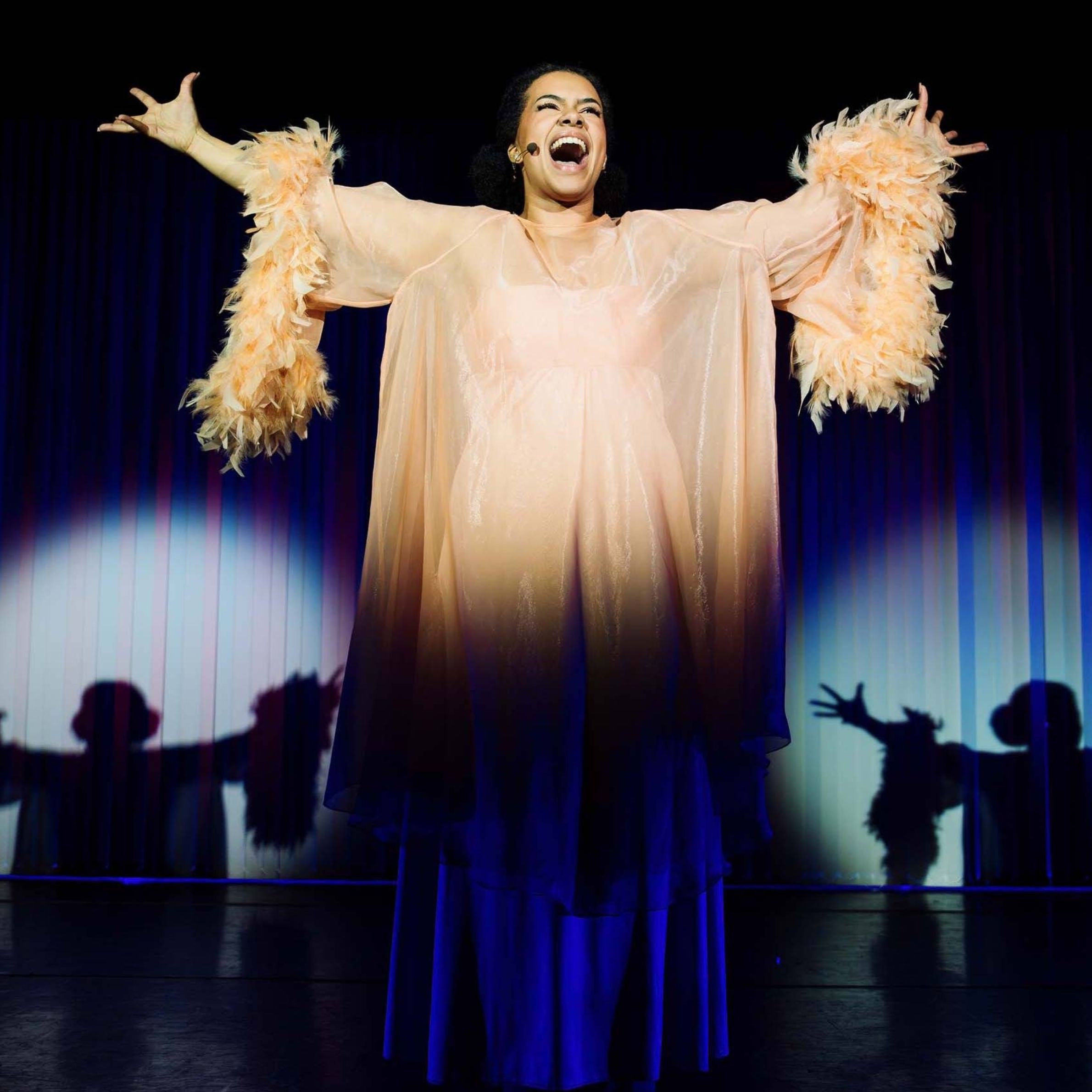 Woman in the centre of a dark stage, lit up with spot light with arms stretch out singing