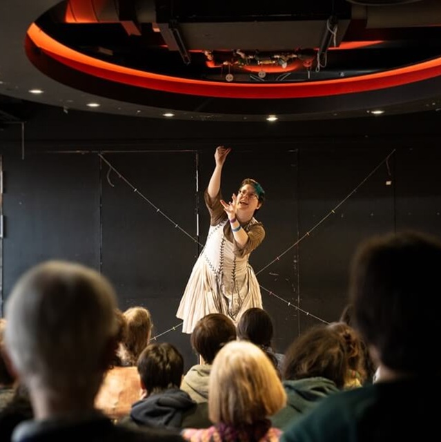 a performer centre stage reaching to the sky, audience members in the foreground