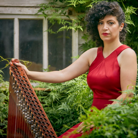 photo of Maya Youssef sat in a garden with lots of green plants and holding her qanun instrument. She is wearing a red dress.
