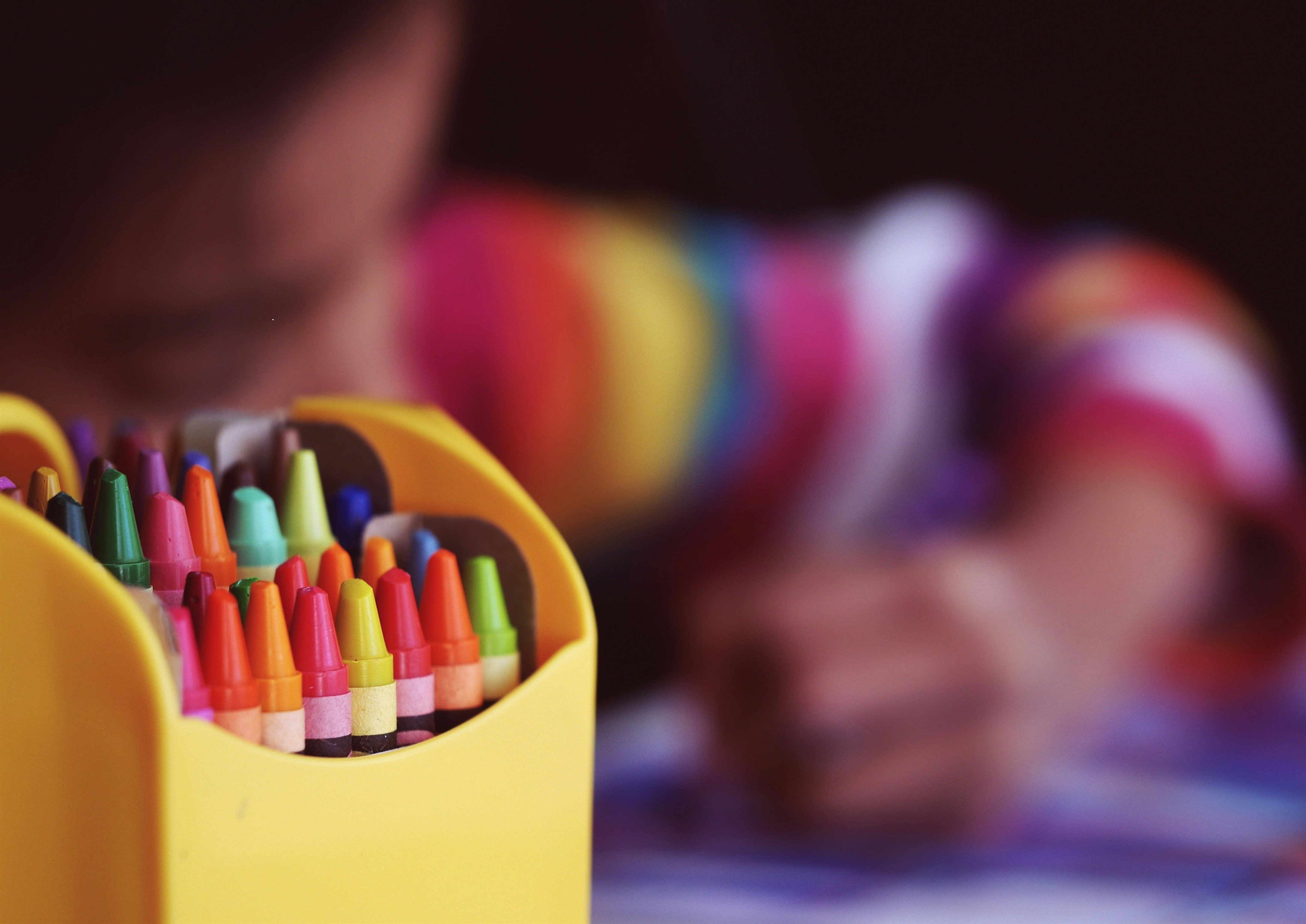 A box of colourful crayons on the left hand side of the picture, a blurred out child is behind.